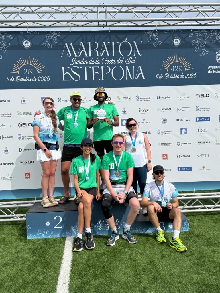 Group of seven runners wearing medals pose on a winners’ podium in front of a sponsor backdrop at the Maratón Estepona event. A large banner above displays “Maratón Jardín de la Costa del Sol Estepona” with race distances of 21K and 42K and the date 11 October 2026. Some participants wear green event shirts, while others wear white and blue race tops. One runner holds a commemorative plaque. The photo was taken outdoors on a sunny day.